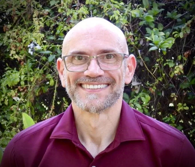 A man with a bald head, glasses, and a beard smiling outdoors in front of green foliage, wearing a maroon shirt.