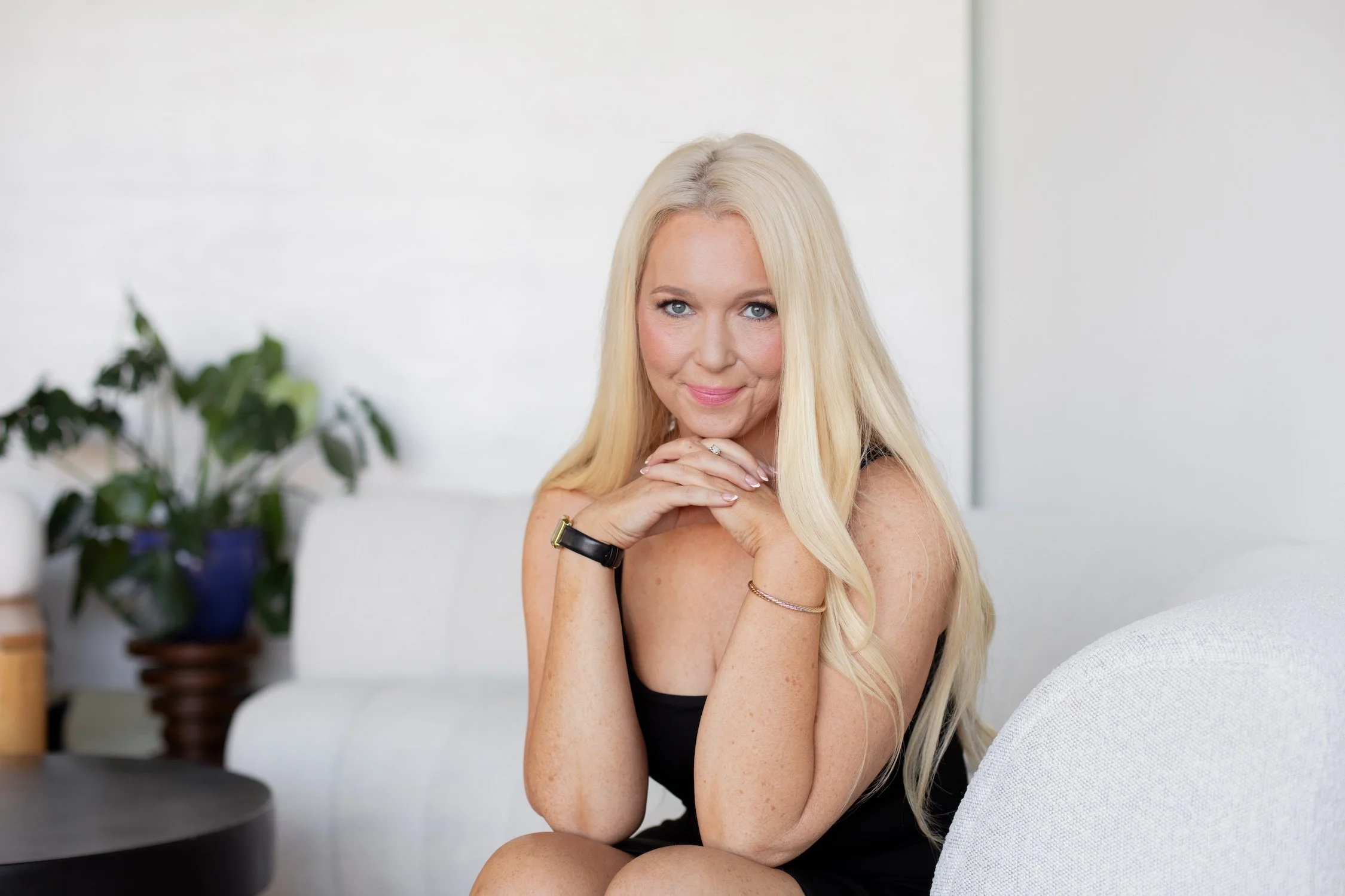 A blonde woman with blue eyes sitting on a white sofa, smiling gently with her hands resting near her chin, in a bright room with a potted plant in the background.