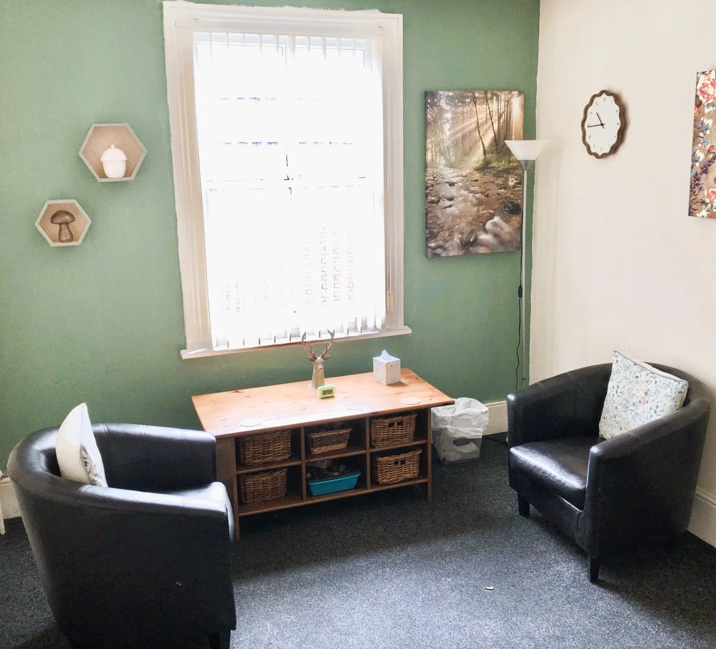 A cozy living room with two black armchairs, a wooden storage unit, and artwork on the walls. There's a large window with white blinds, a standing lamp, and decorative items like a deer head sculpture, a clock, and a framed nature photo.