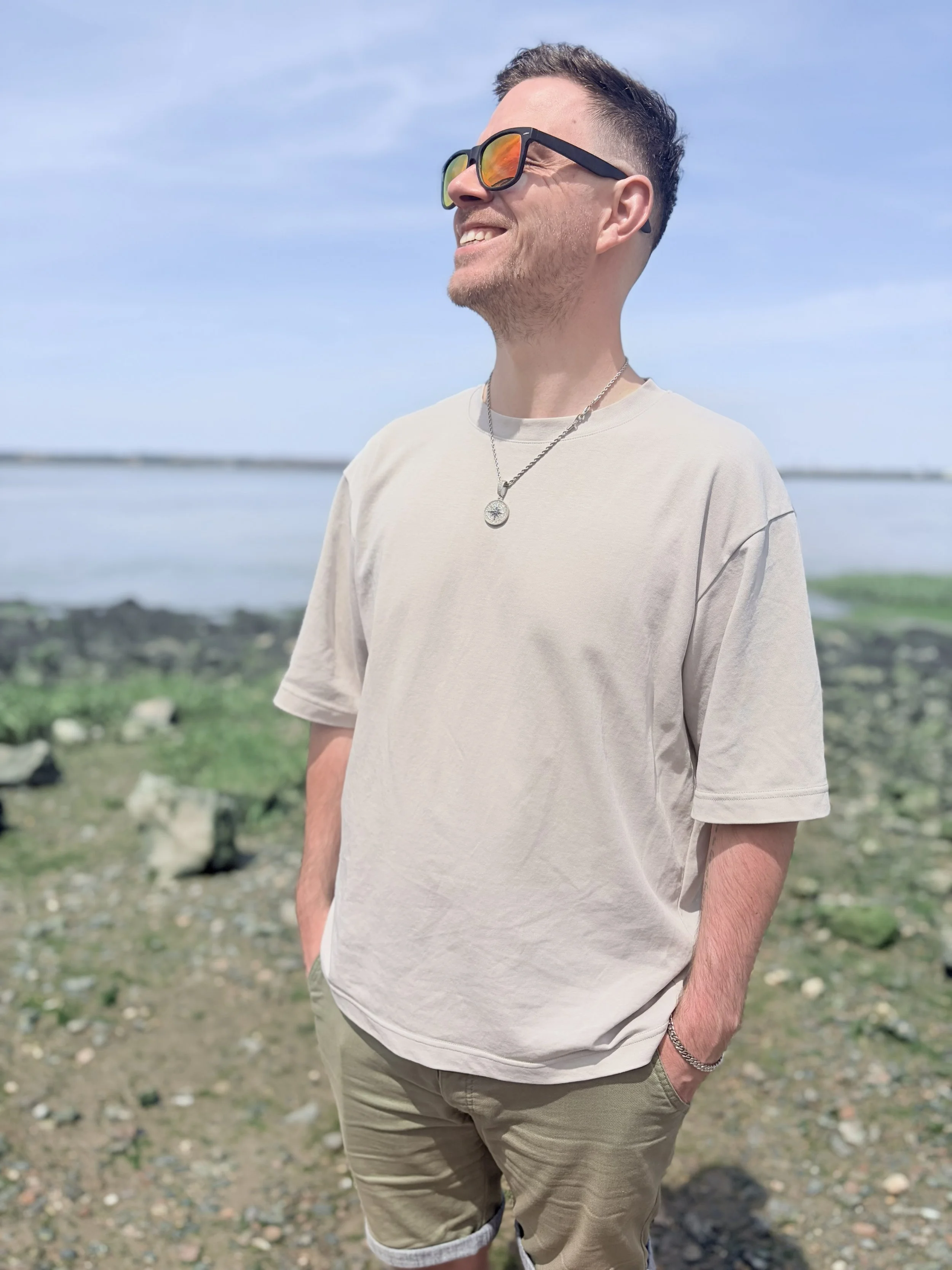 A man standing outdoors near a body of water, smiling, wearing sunglasses, a beige T-shirt, khaki shorts, and a silver necklace.