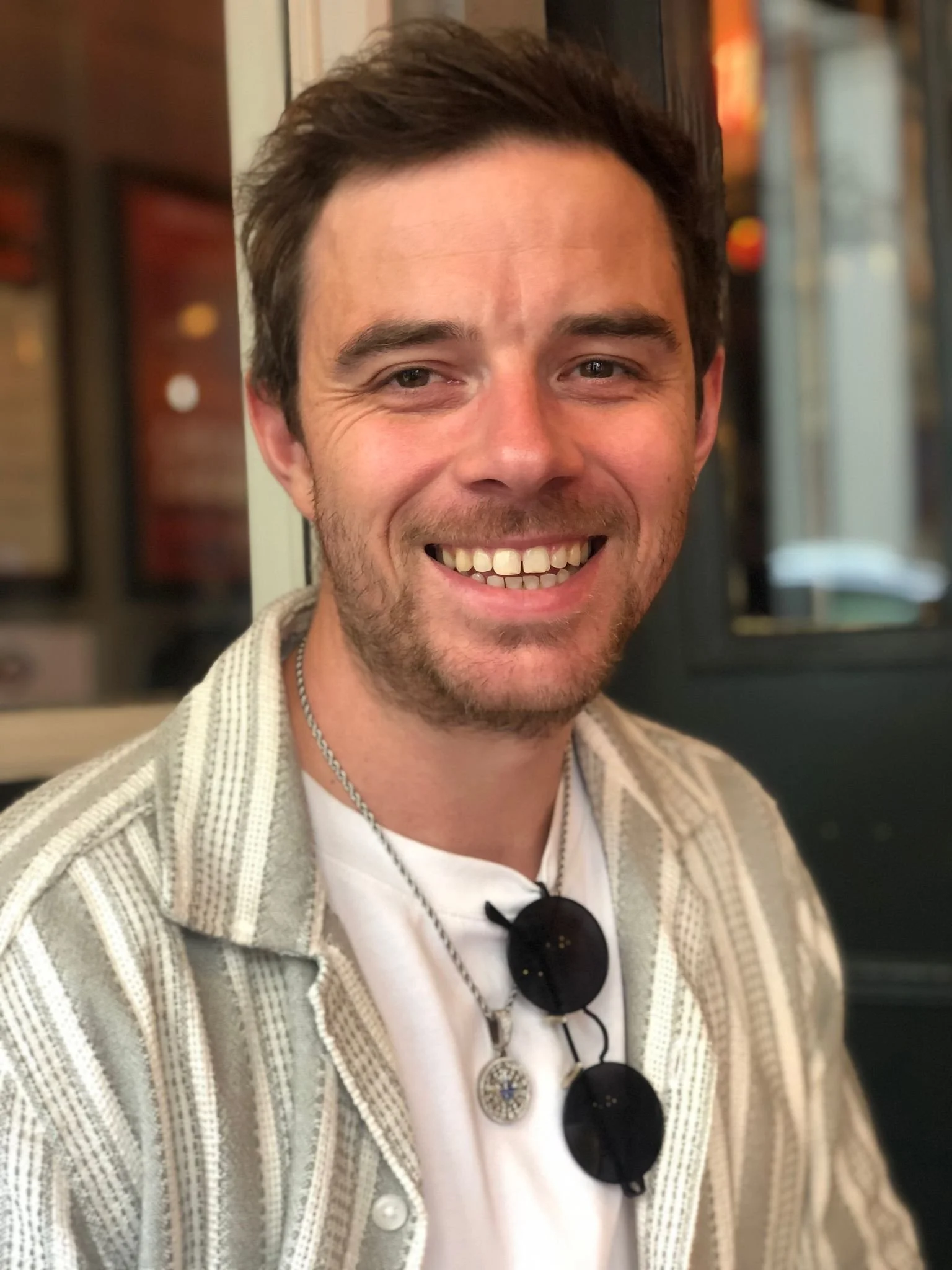 Portrait of a smiling man with short dark hair and facial hair, dressed in a white shirt and a beige striped cardigan, wearing black sunglasses hanging around his neck, in an indoor setting with blurred background.