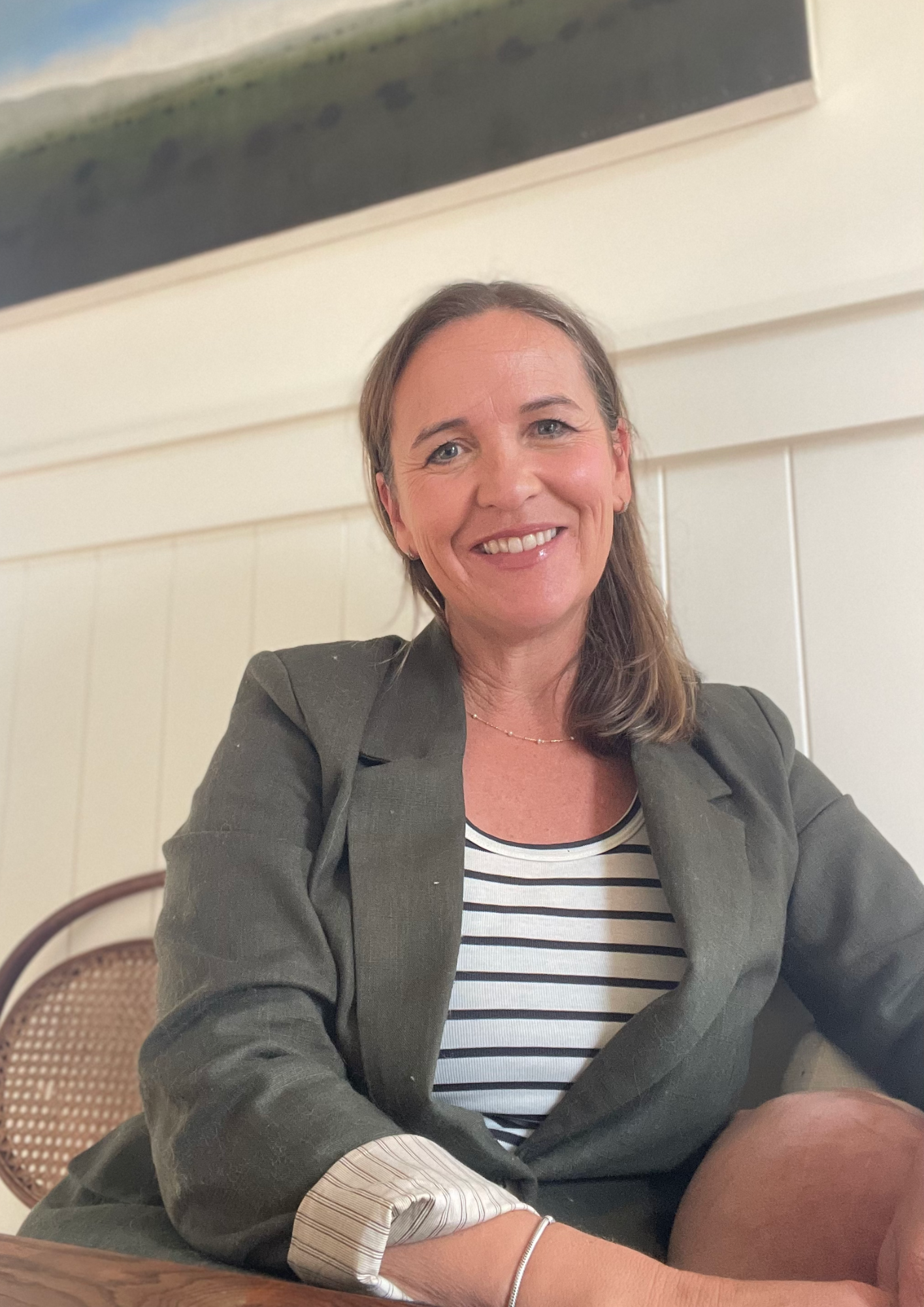 A woman with shoulder-length brown hair smiling, wearing a gray blazer over a striped shirt, sitting at a wooden table, with a cream paneled wall behind her and a piece of artwork above.