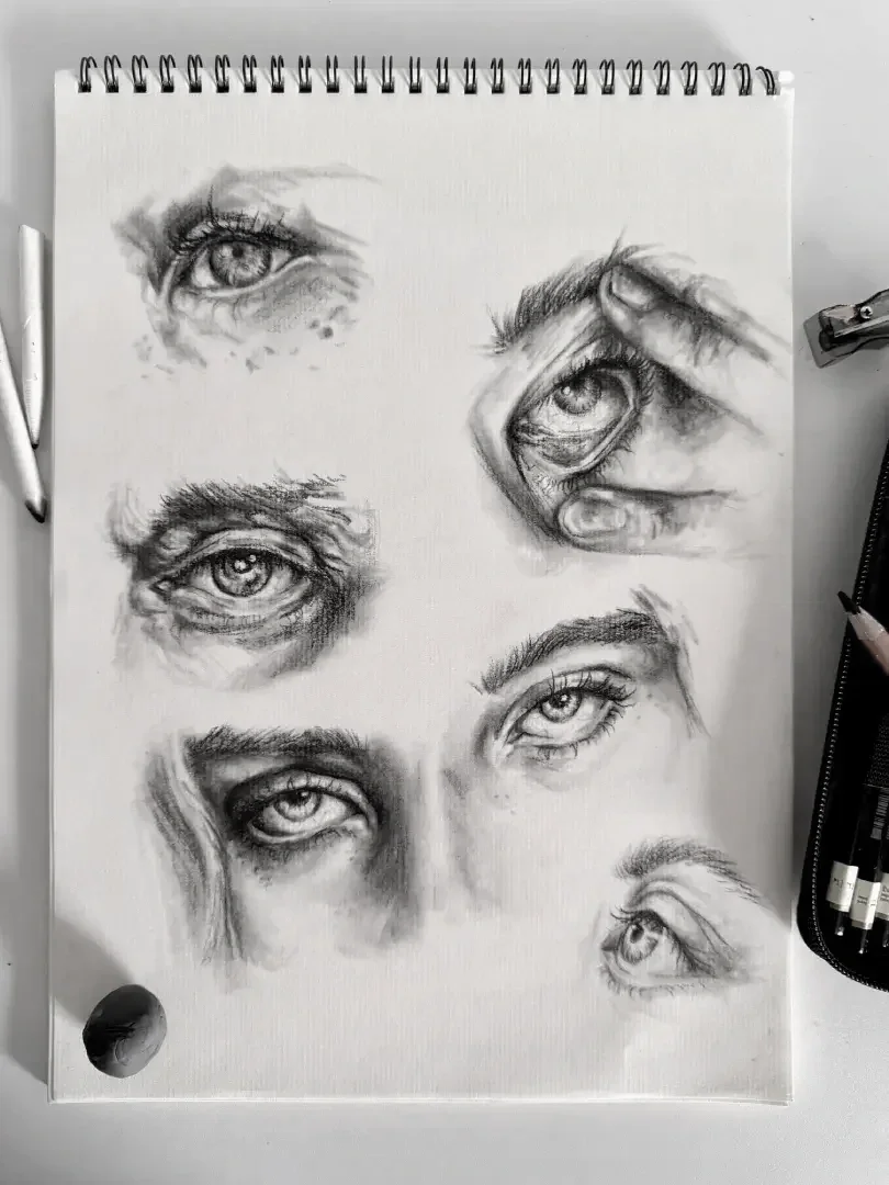 A sketchbook page displaying five detailed graphite pencil sketches of human eyes, along with an eye partially covered by a hand, showcasing various expressions and angles.