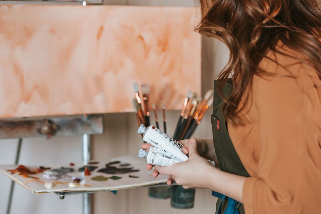 A woman holding tubes of paint and brushes in an art studio or workspace.