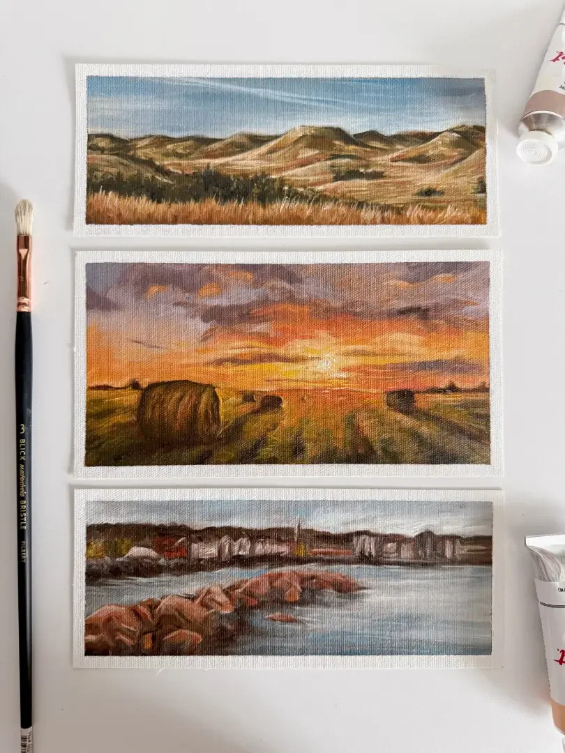 Three landscape paintings on paper, depicting a mountain scene, a sunset over a field, and a rocky shoreline with water in the foreground, placed on a white surface alongside a paintbrush and tubes of paint.