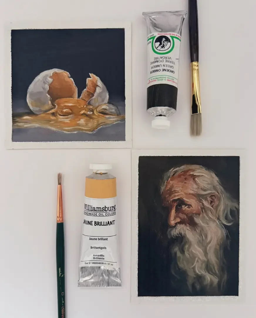 Two small watercolor paintings, one of a baby emerging from an eggshell with a spilled yolk, and the other of a bearded elderly man with a serious expression. The paintings are accompanied by two paintbrushes and two tubes of oil paint, one of which 