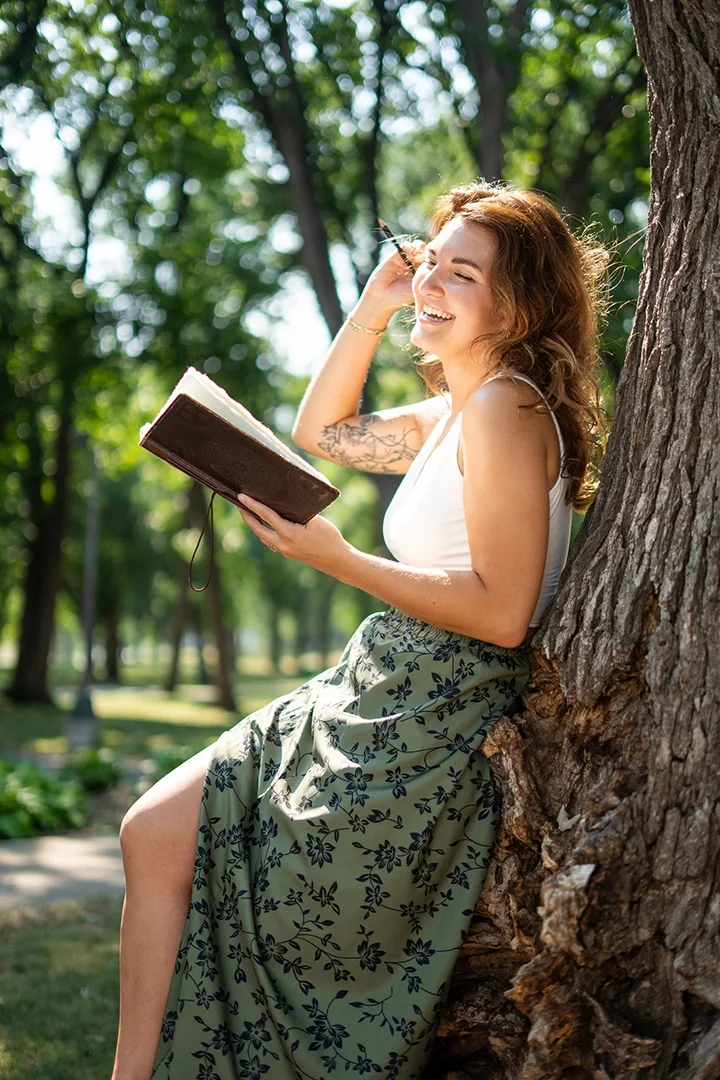 A woman with curly brown hair sitting against a tree, smiling, reading a book in a park with green trees in the background.