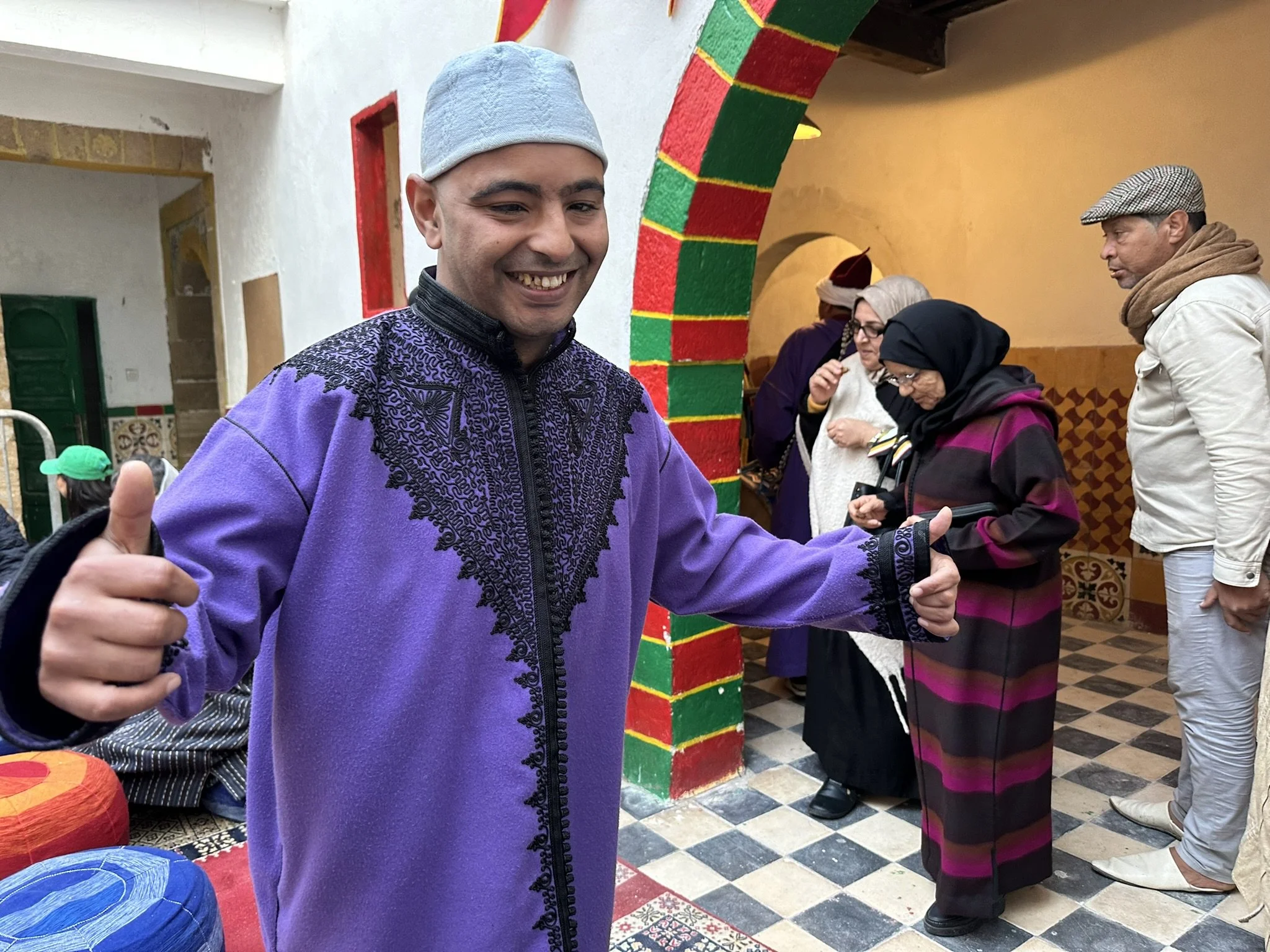 A smiling man in a purple embroidered traditional outfit and gray cap, giving a thumbs-up, at a colorful indoor cultural gathering with other people and vibrant decorations.