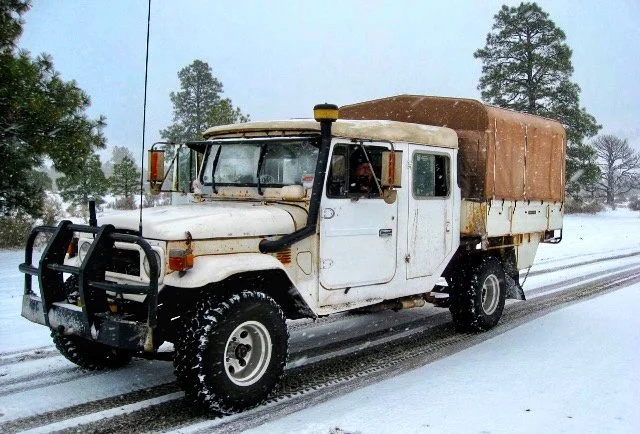 An old 1983 Land Cruiser truck with a tan cargo cover driving on a snowy road with trees in the background.