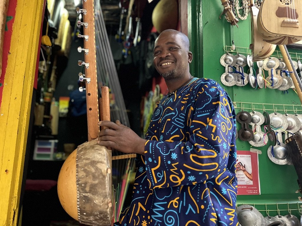 A man smiling and holding a traditional musical instrument in a shop with hanging steel cups and musical gear.