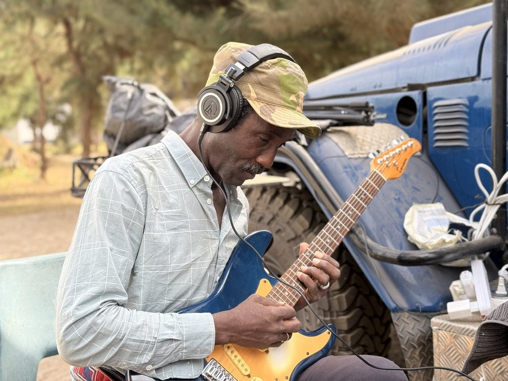 A man wearing a camouflage hat, headphones, and a light plaid shirt playing a blue electric guitar outdoors near a blue off-road vehicle.