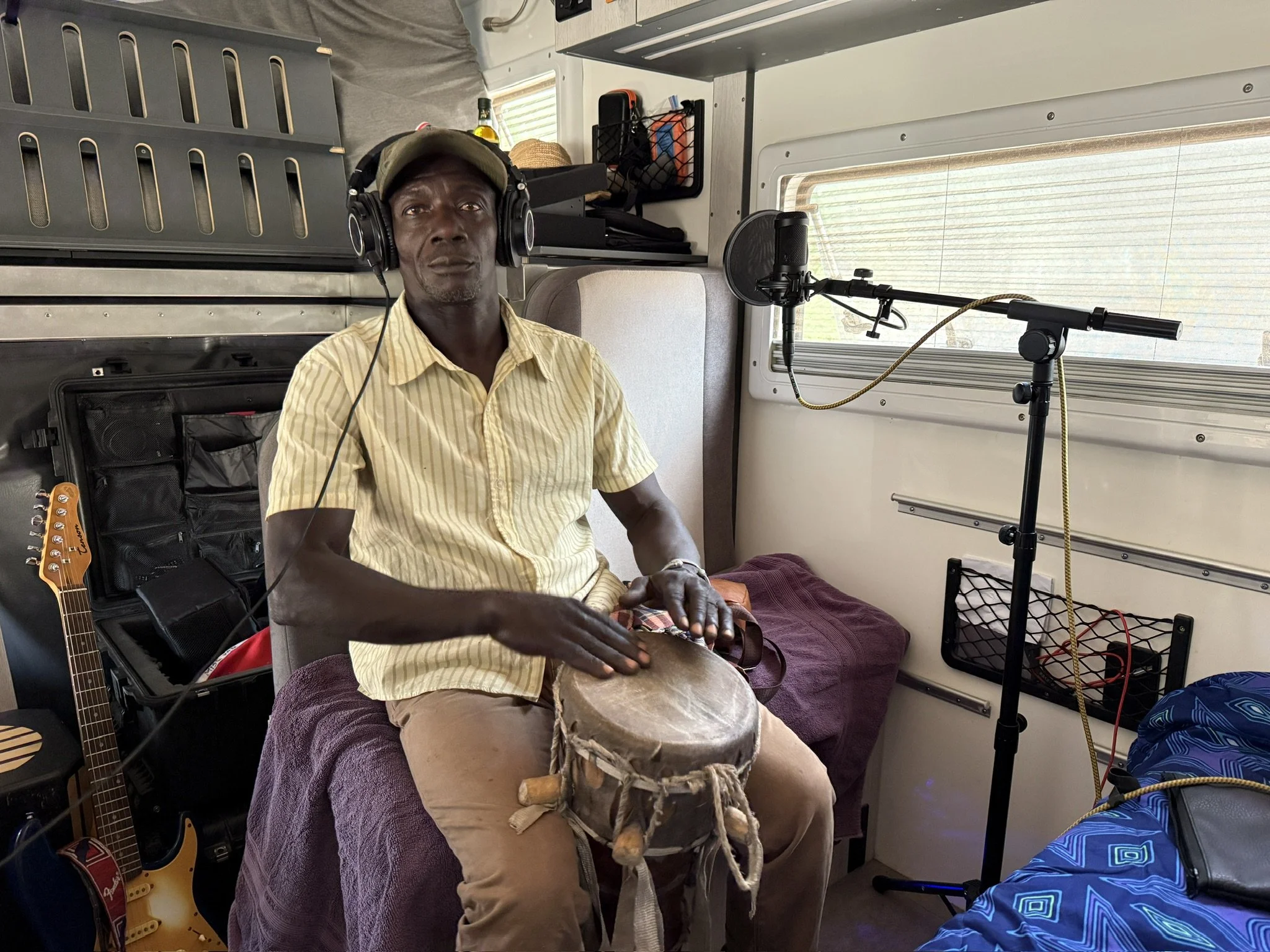 A man wearing a tan cap and yellow striped shirt sitting inside a vehicle with a drum on his lap, a microphone on a stand nearby, and musical instruments and equipment in the background.