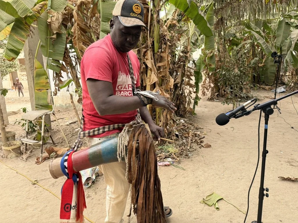 Man in a red shirt and beige pants playing a didgeridoo outdoors near banana trees with a microphone set up nearby.