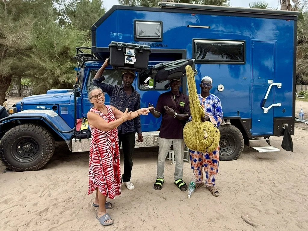 Four people standing outdoors in front of a large blue camper truck, with trees in the background. One woman is wearing a red and white patterned dress, smiling and pointing. The other three men are dressed casually, one holding a yellow guitar-shape