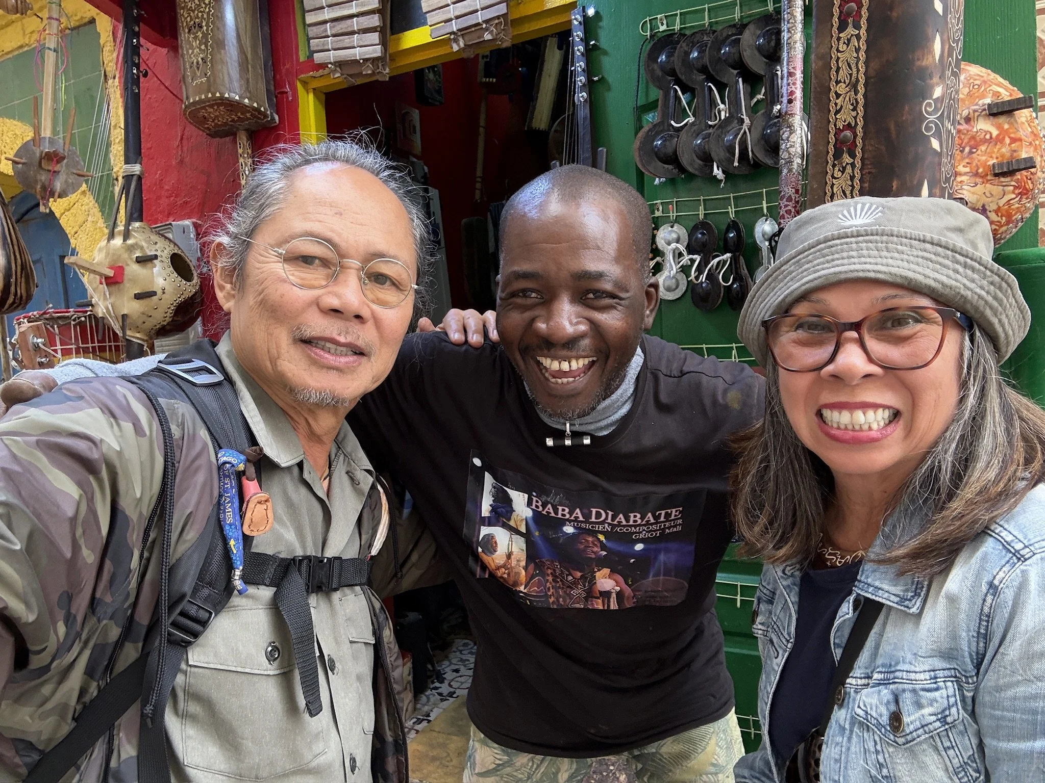 Three smiling people, two women and a man, are taking a selfie in front of a marketplace stall. The stall displays various traditional musical instruments, including drums and balafons, and some artwork. The individuals are wearing casual clothing an