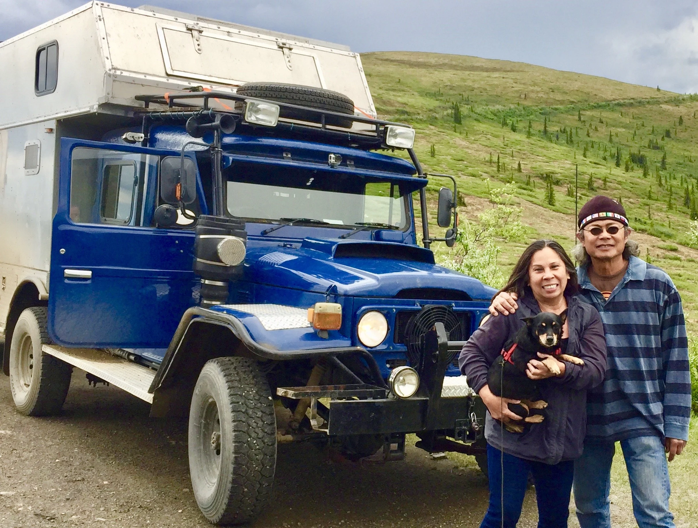 Two people, a woman and a man, standing outside near a 1983 Land Cruiser with a camper attached, holding a small black dog. The background features a grassy hillside with scattered trees and a cloudy sky.