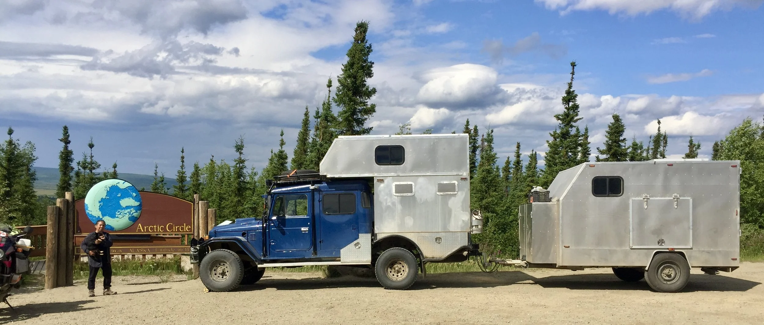 A blue truck with a large silver camper attached, parked near a sign that reads 'Arctic Circle' with a globe illustration. There are trees, mountains, and a partly cloudy sky in the background. A person is standing near the sign.