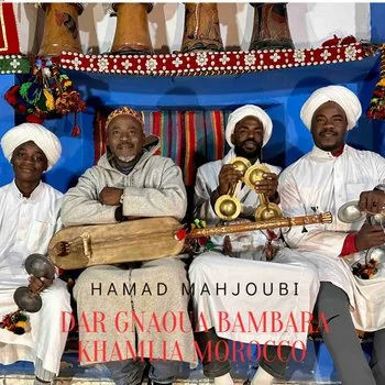 Four Gnaouan men dressed in traditional North African attire sitting on a decorated stage, with two men holding musical instruments, one with a large drum and another with a stringed instrument called a guembri, against a colorful backdrop.