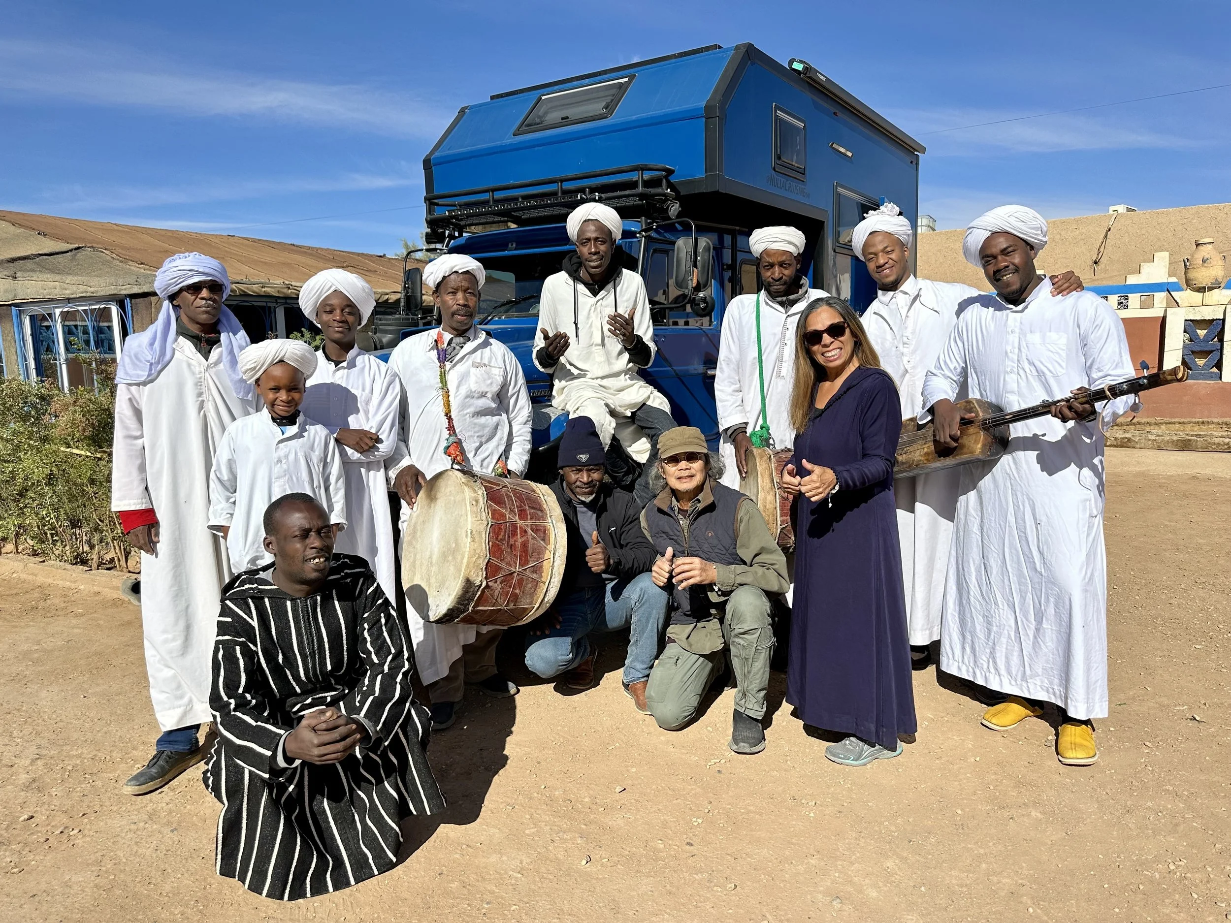 A group of people, including men, women, and children, dressed in white traditional clothing and head coverings, standing outdoors in front of a blue camper Land Cruiser truck and a rural setting with some simple buildings and a clear blue sky.