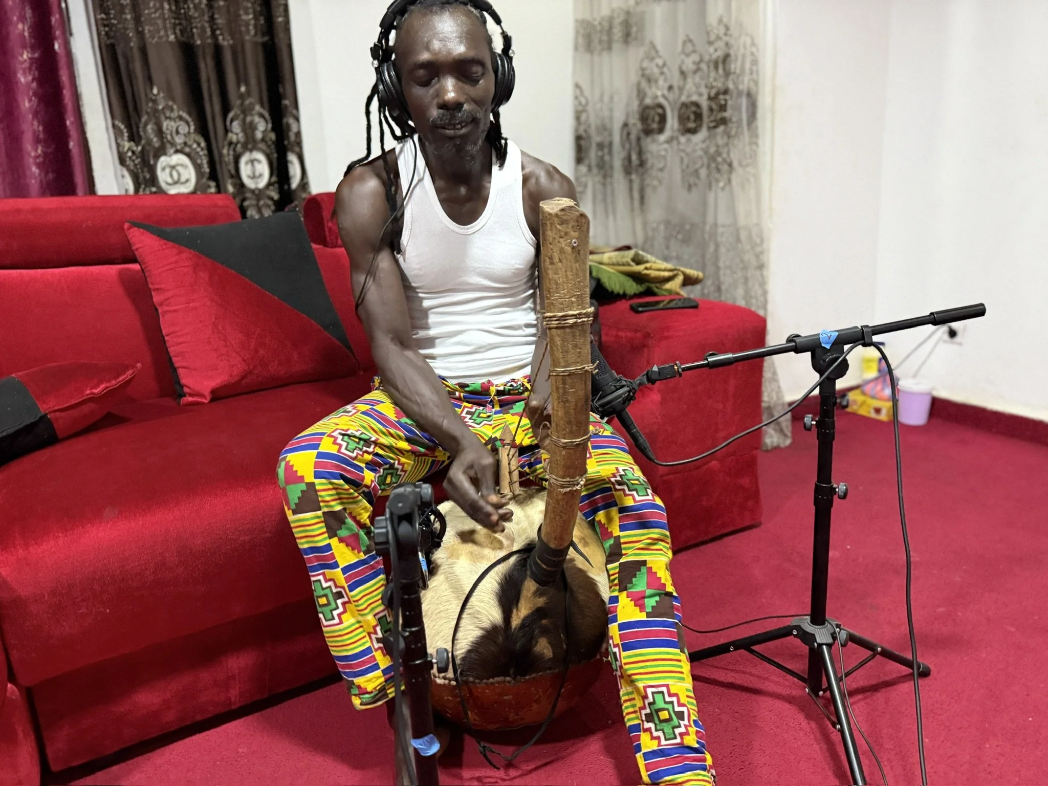 An elderly man with dreadlocks wearing a white tank top and colorful patterned pants, sitting on a red sofa, playing a traditional wooden musical instrument with a microphone set up in front of him.