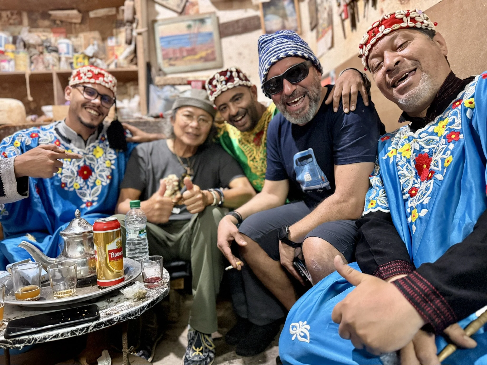 Group of six people smiling and enjoying drinks at a gathering. They are wearing colorful traditional clothing and some have headgear made of shells. The scene appears to be inside a rustic, cozy room with shelves and paintings in the background. The