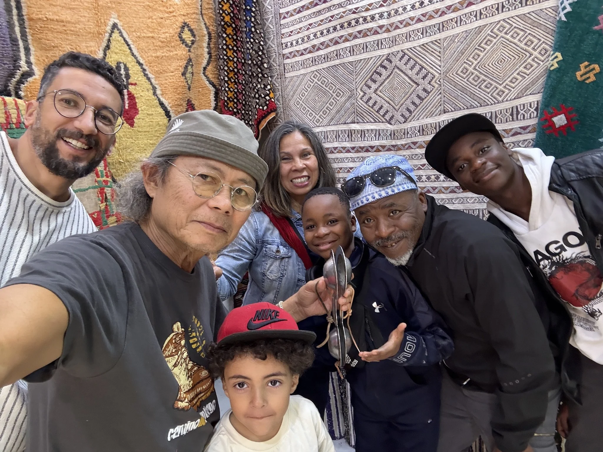 A group of diverse people, including two children and adults, smiling and posing for a selfie in front of decorative Moroccan woven textiles hanging on a wall.