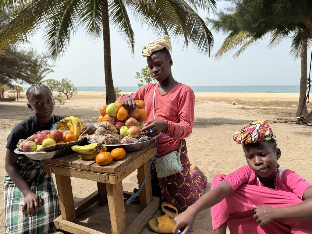 Three children selling fruits at a beachside market with palm trees in the background.