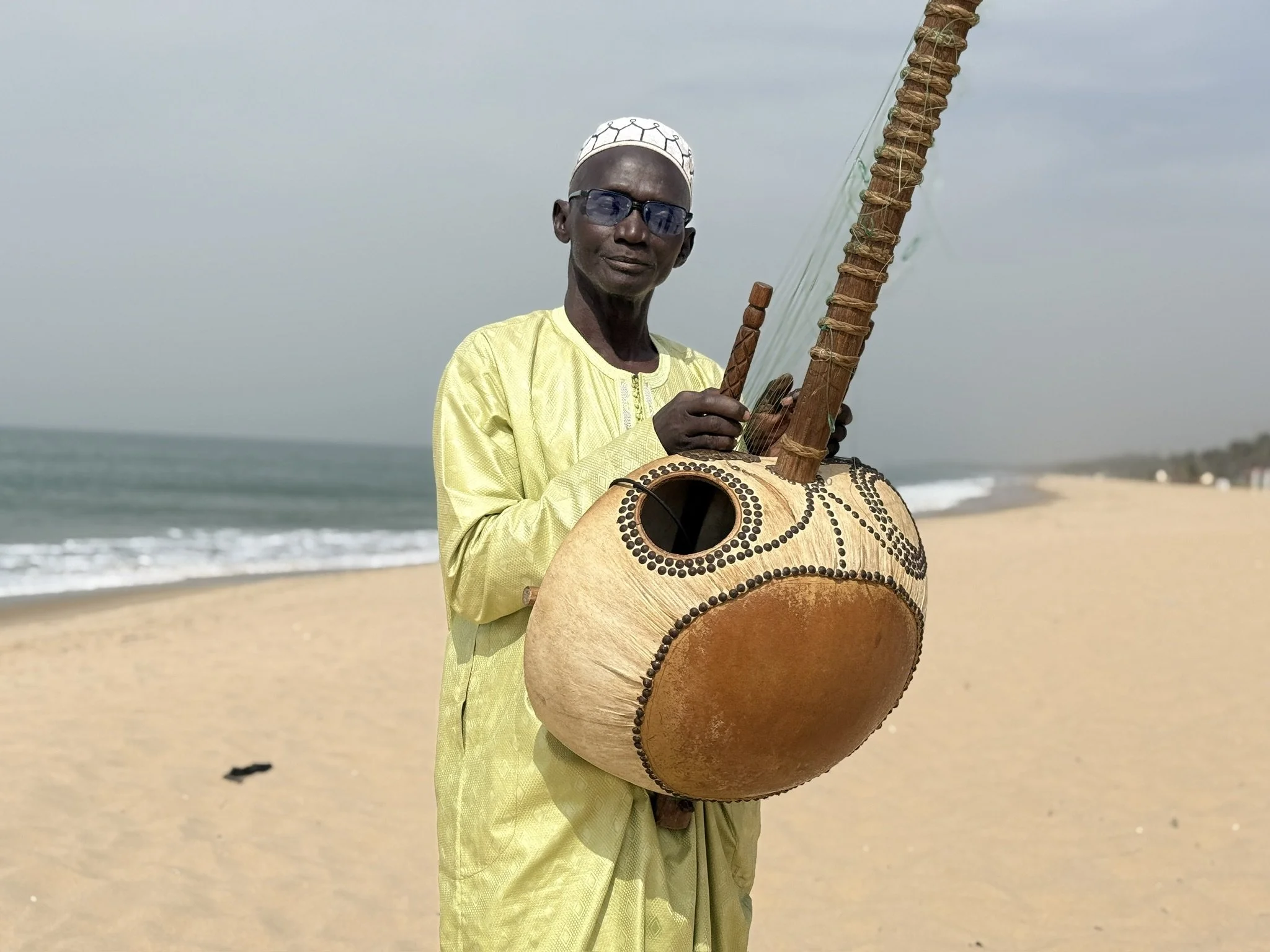 A man in yellow attire wearing glasses and a white cap, holding a large wooden drum with decorative dots, standing on a beach near the ocean with a cloudy sky.