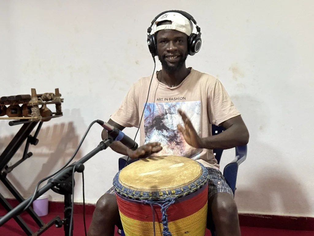 A man wearing a beige t-shirt, plaid shorts, headphones, and a backward cap playing a drum.