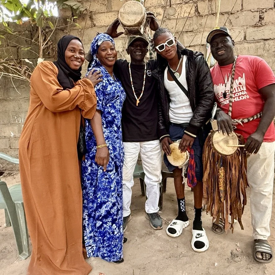 Group of five people smiling, standing outdoors in front of a brick wall, with musical drums and one person playing traditional African instrument.