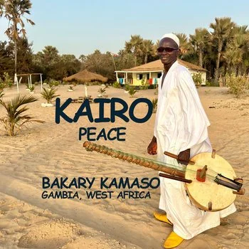 A man dressed in white traditional Gambian clothing and yellow shoes holding a large stringed instrument, standing on a sandy area with palm trees and a building in the background. Text reads: 'KAIR0 PEACE BAKARY KAMASO GAMBIA, WEST AFRICA.'
