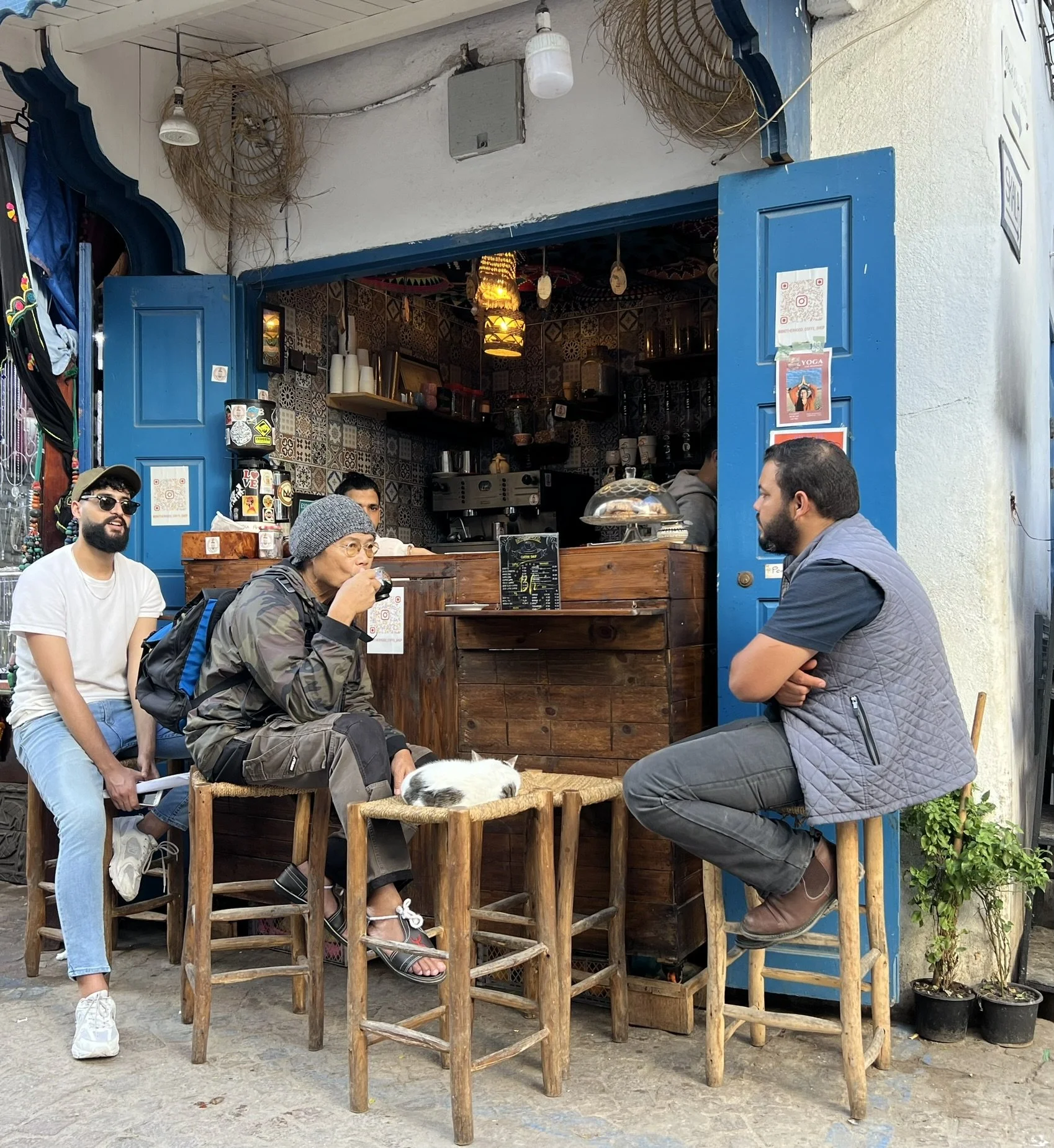People sitting and standing outside a small cafe or restaurant with blue doors and indoor decor. A dog is resting on a stool in the foreground.