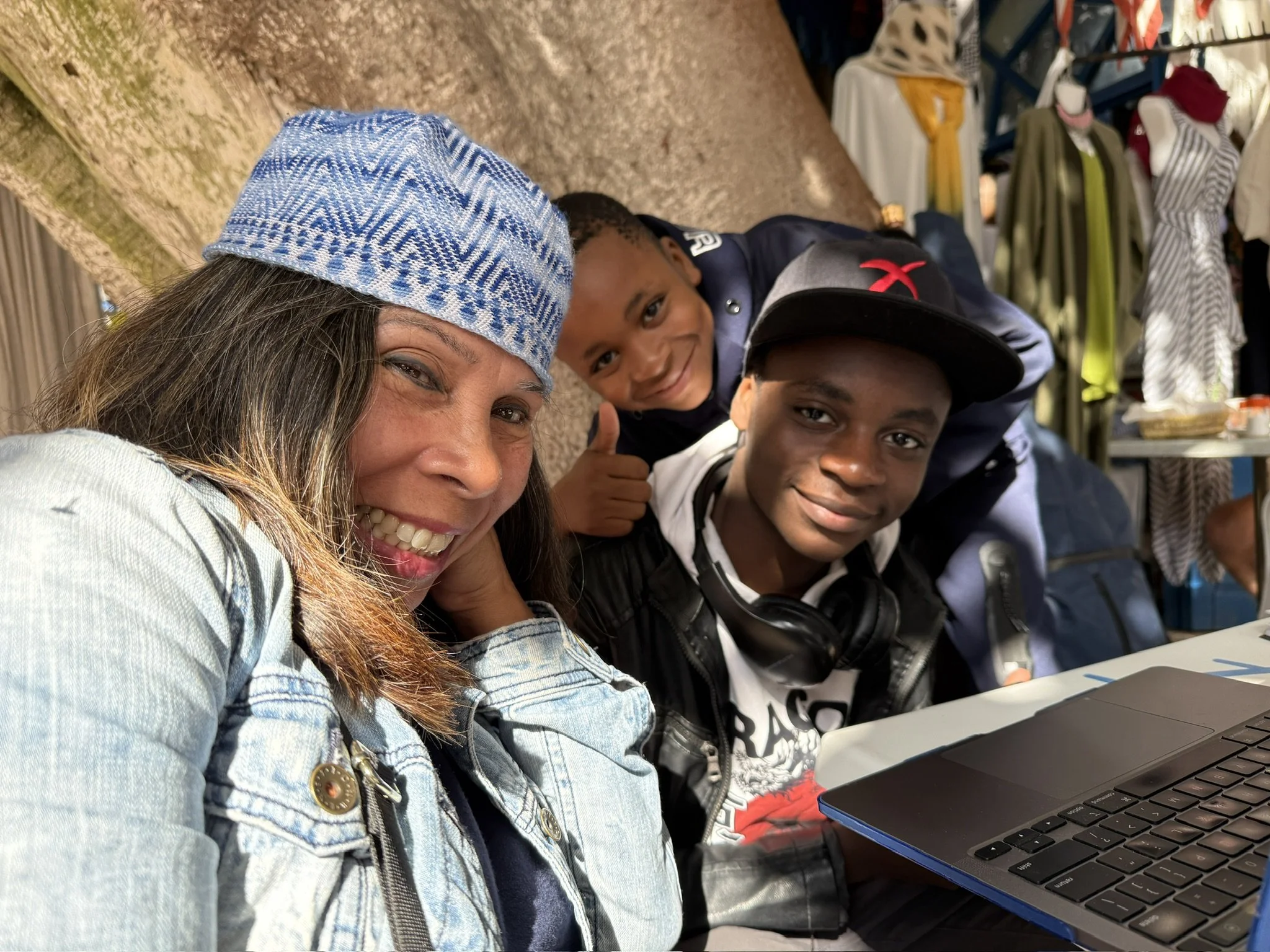 A woman with long hair wearing a blue patterned headscarf and denim jacket smiling at the camera, with two young boys behind her giving thumbs up, in a room with clothes on racks and a laptop on the table.