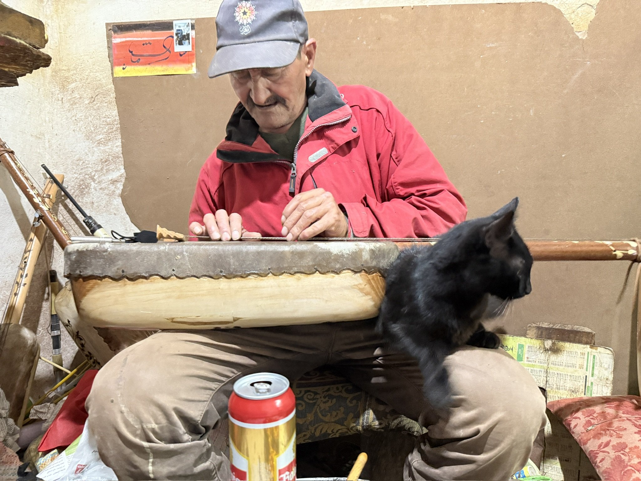An elderly man wearing a gray cap and red jacket sits at a table, playing a stringed musical instrument. A black dog is sitting on the floor nearby. The background features a wall with peeling paint and a colorful sign with Arabic writing.