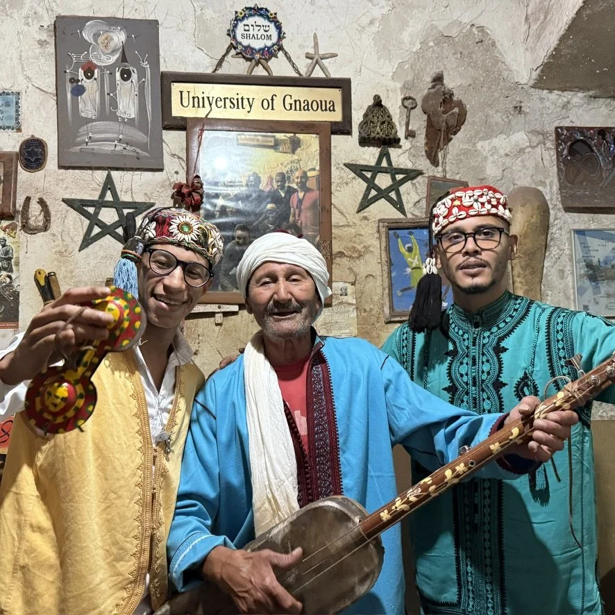 Three men dressed in traditional Moroccan attire posing with musical instruments in a decorated room. The background includes various artwork, symbols, and signs, including one that reads "University of Gnaoua".