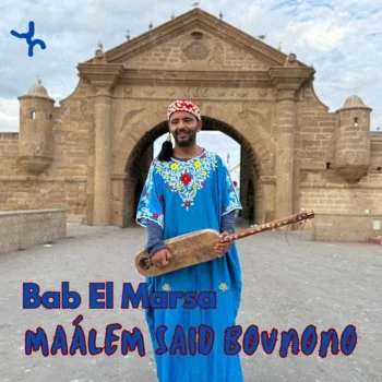 Man dressed in traditional Moroccan blue attire with embroidery, holding a musical instrument called a guembri in front of an historic stone gateway.