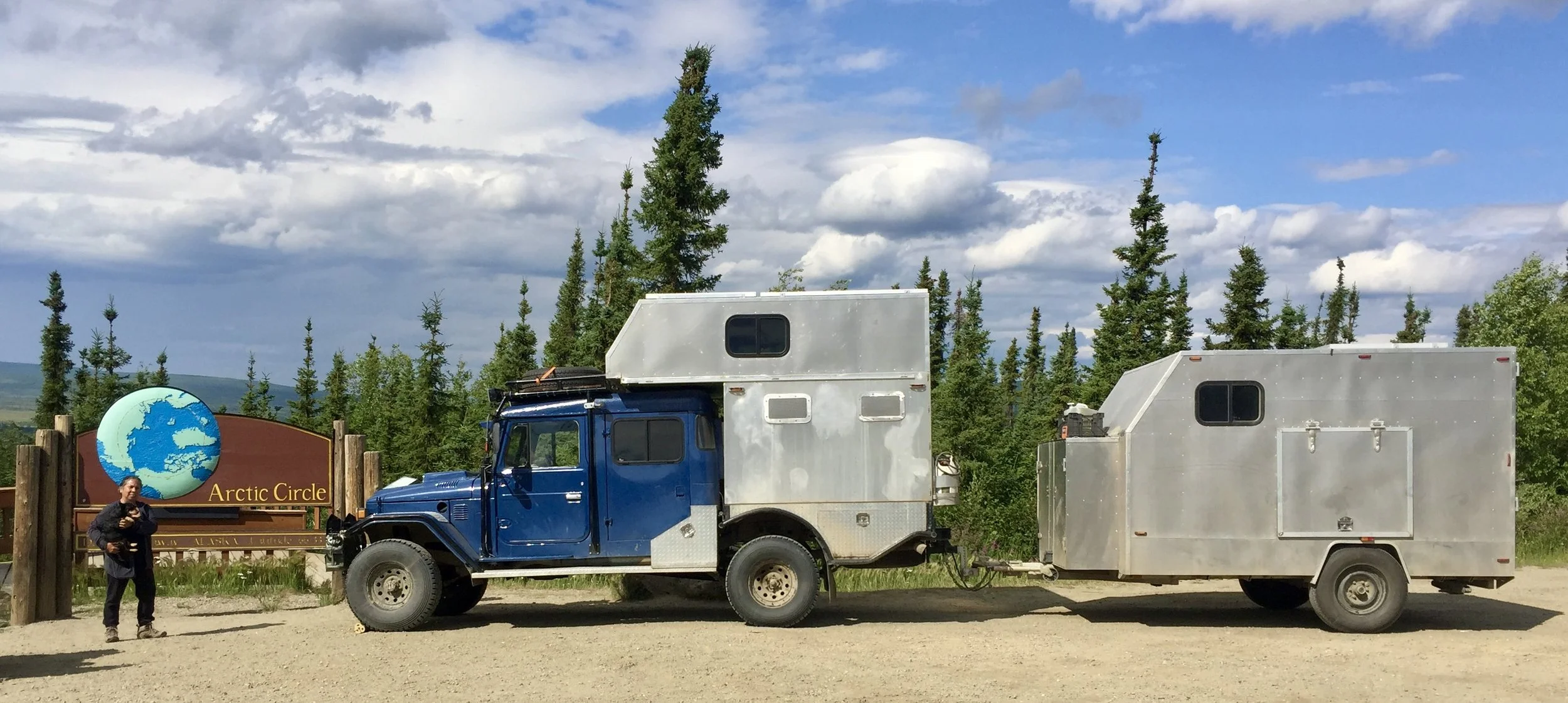 A person standing near a sign that reads 'Arctic Circle' and a large blue Land Cruiser truck with a silver camper and trailer, with a forest of evergreen trees and a partly cloudy sky in the background.