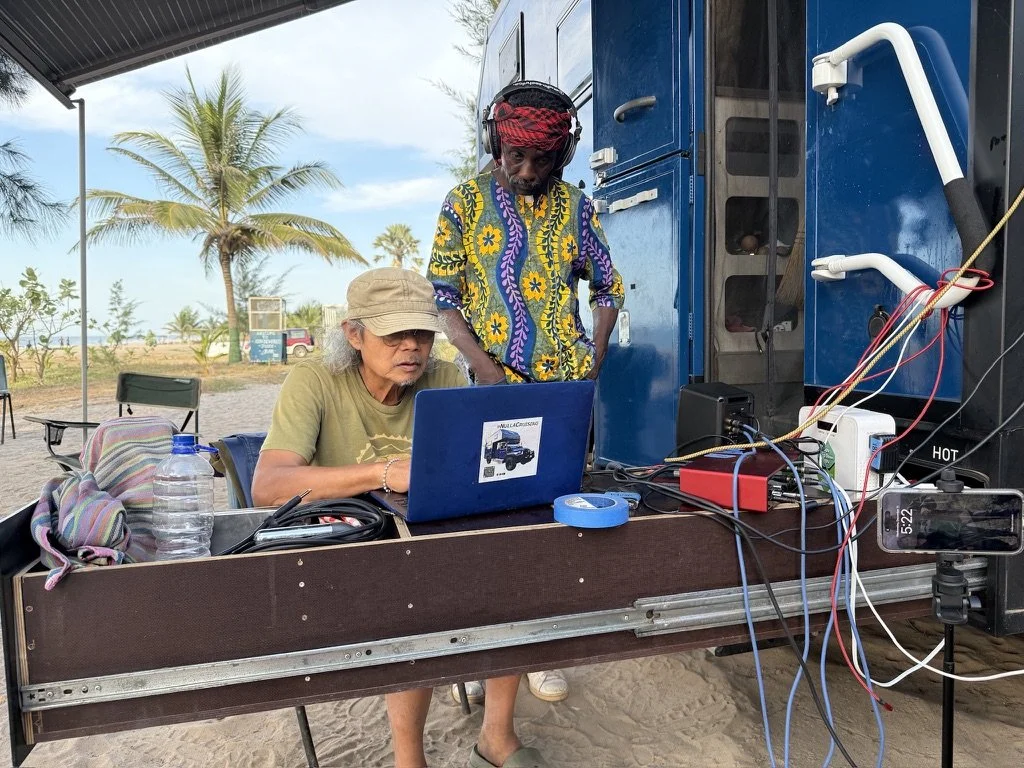 Two people working with a laptop at an outdoor setup, with palm trees in the background. One woman wearing a hat, and a man wearing a colorful shirt and headphones.