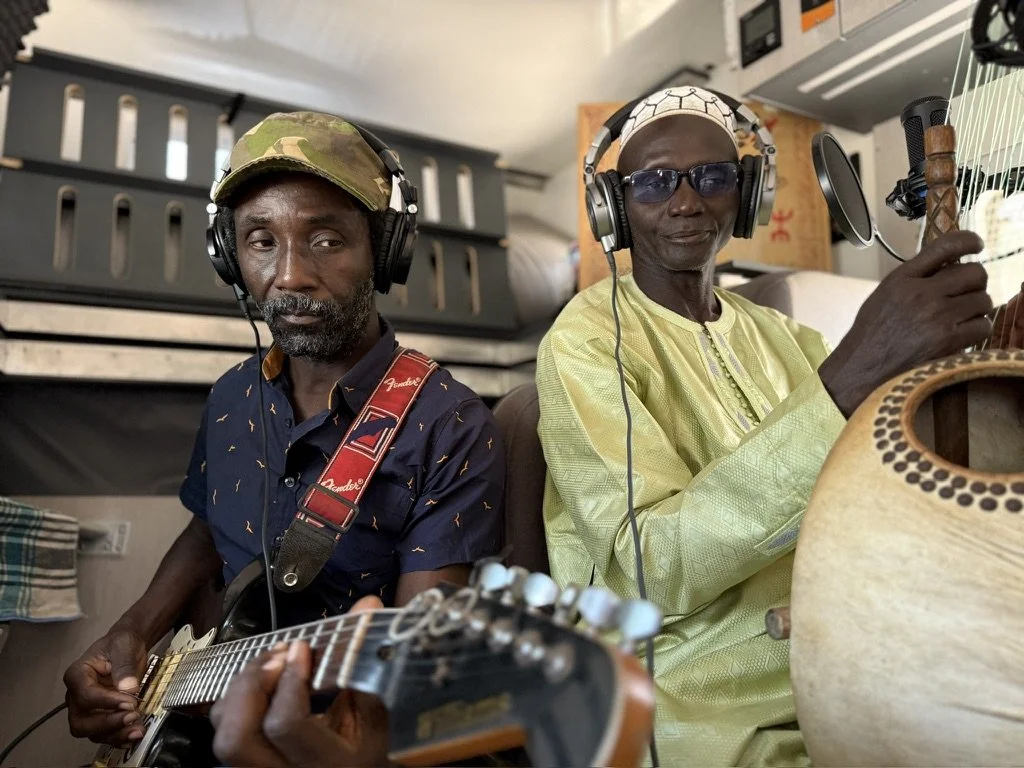 Two men wearing headphones, one playing an electric guitar and the other playing a traditional Kora instrument, inside a room.