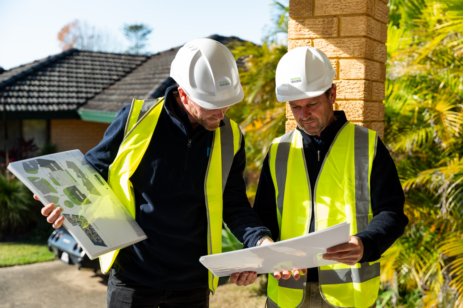 Insurance builders in Adelaide wearing yellow safety vests and white hard hats reviewing blueprints outside near a house with tropical plants in the background.
