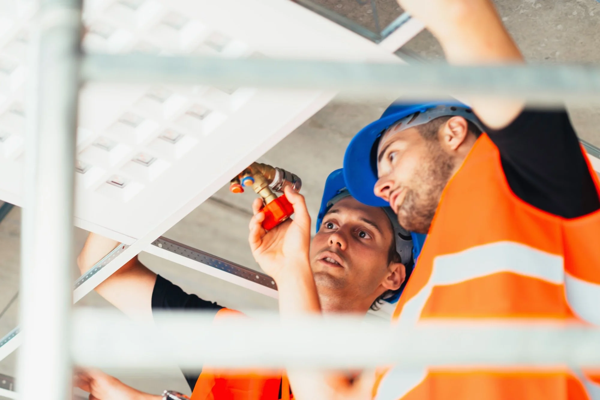 Two construction workers, wearing blue safety helmets and orange safety vests, working on an electrical panel or control box while lying on the floor | Adelaide Insurance Builders | SA