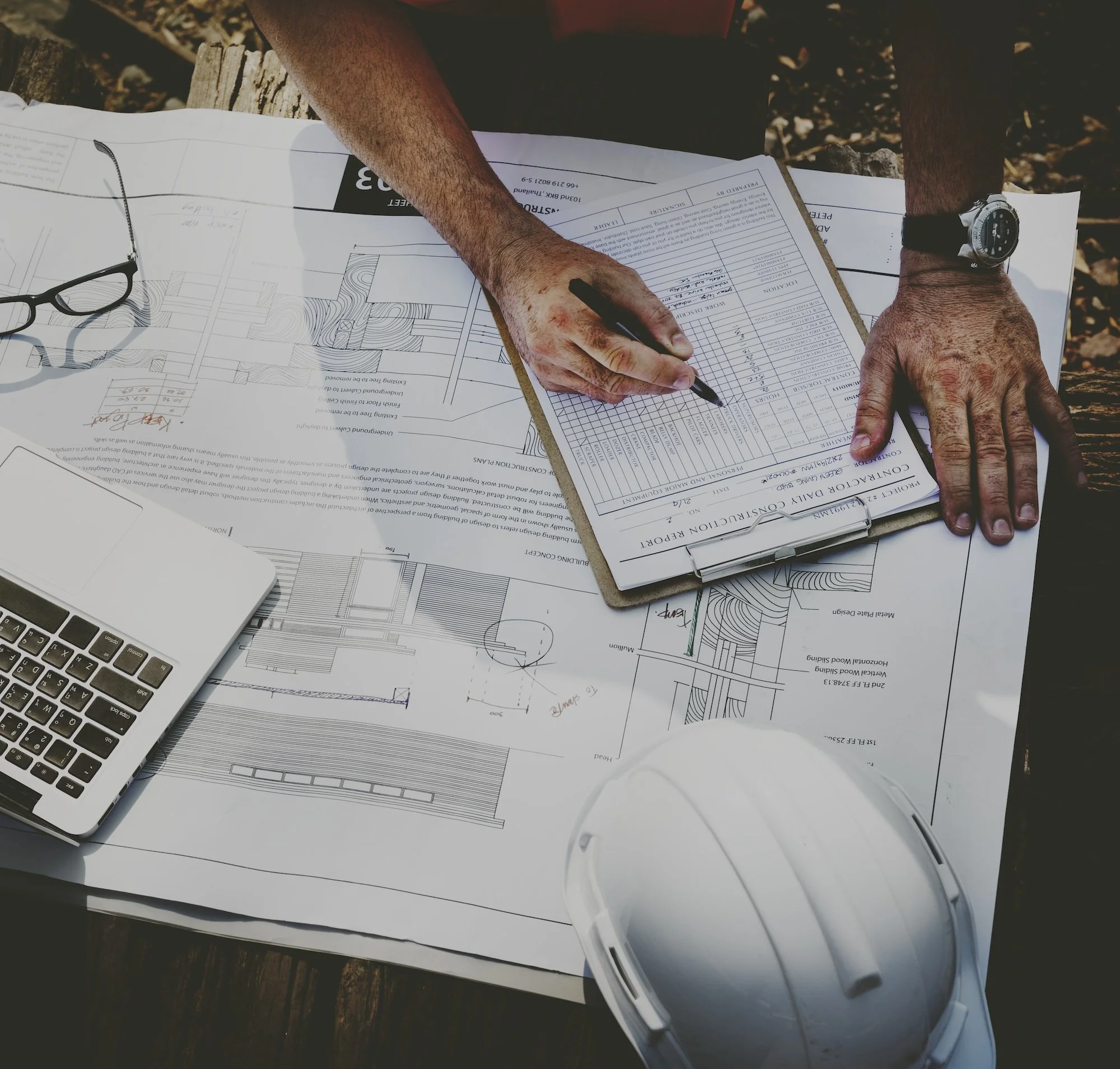 Top-down view of an architectural drafting table with blueprints, a pair of glasses, a laptop, a clipboard with construction documents, a wristwatch, and white rollers, all set on a wooden surface | Adelaide Insurance Builders | SA