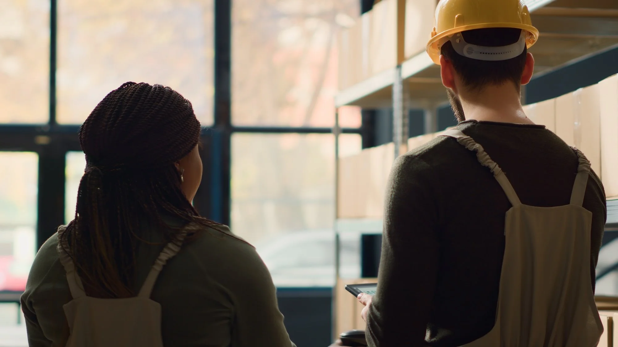 Two people, a woman and a man, are standing indoors near large windows during daylight. The woman has braided hair and is wearing a knit hat and a beige jacket | Adelaide Insurance Builders | SA