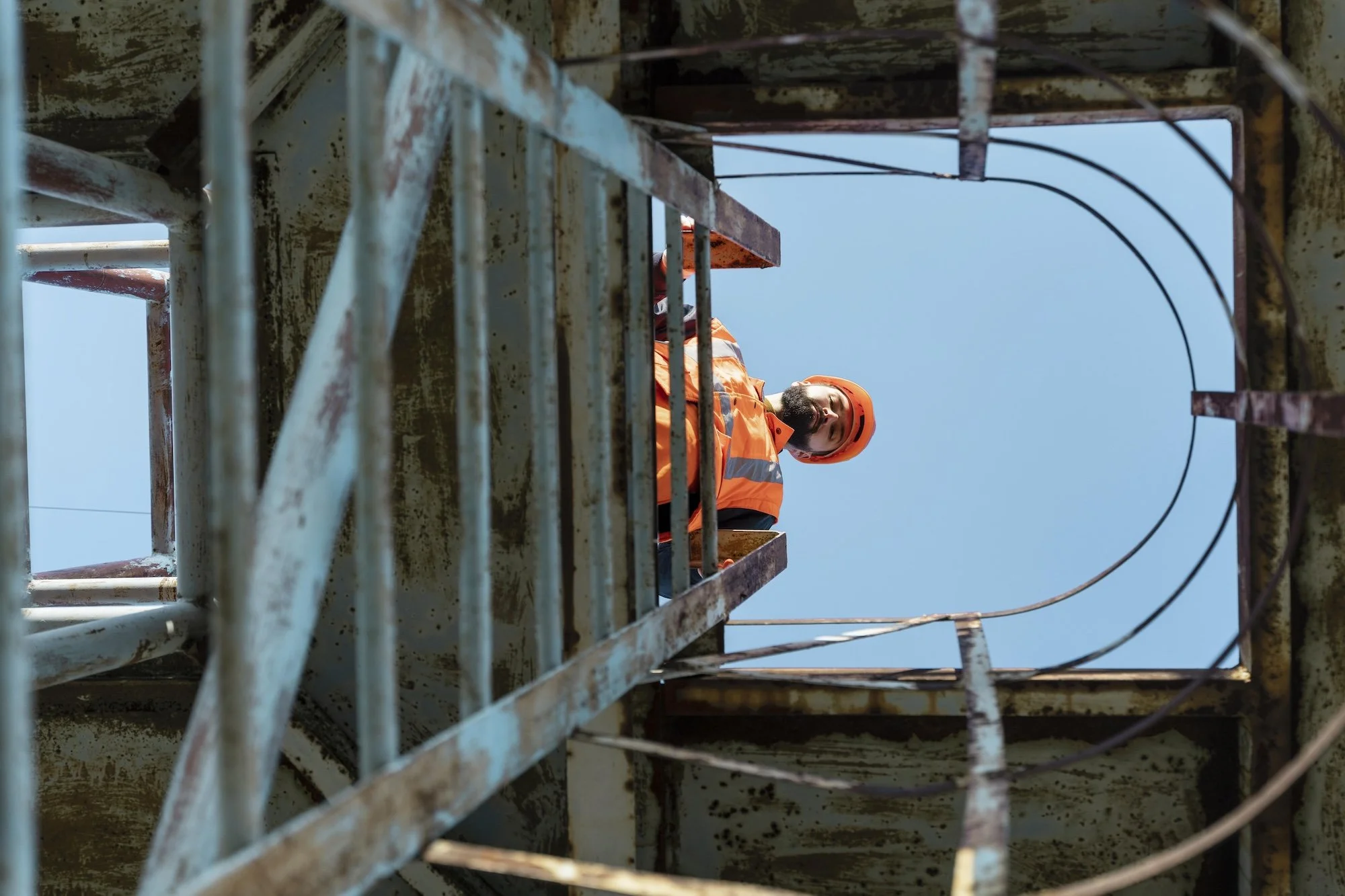 A worker in an orange safety uniform and helmet standing on a rusty steel structure against a clear blue sky | Adelaide Insurance Builders | SA