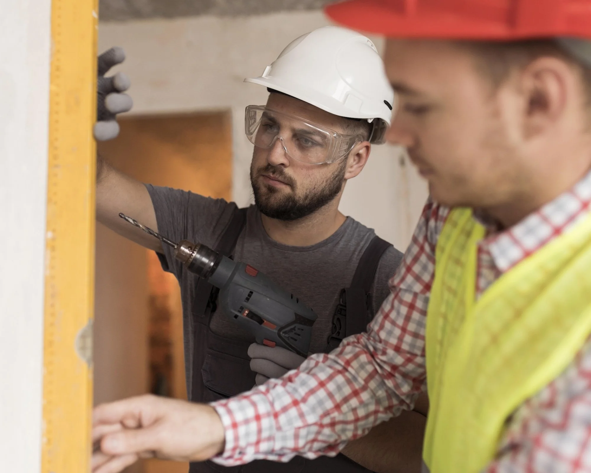 Two construction workers, one in a gray shirt and the other in a plaid shirt and yellow safety vest, working together with tools. One wears a white safety helmet and safety glasses, and the other wears a red safety helmet | Adelaide Insurance Builder