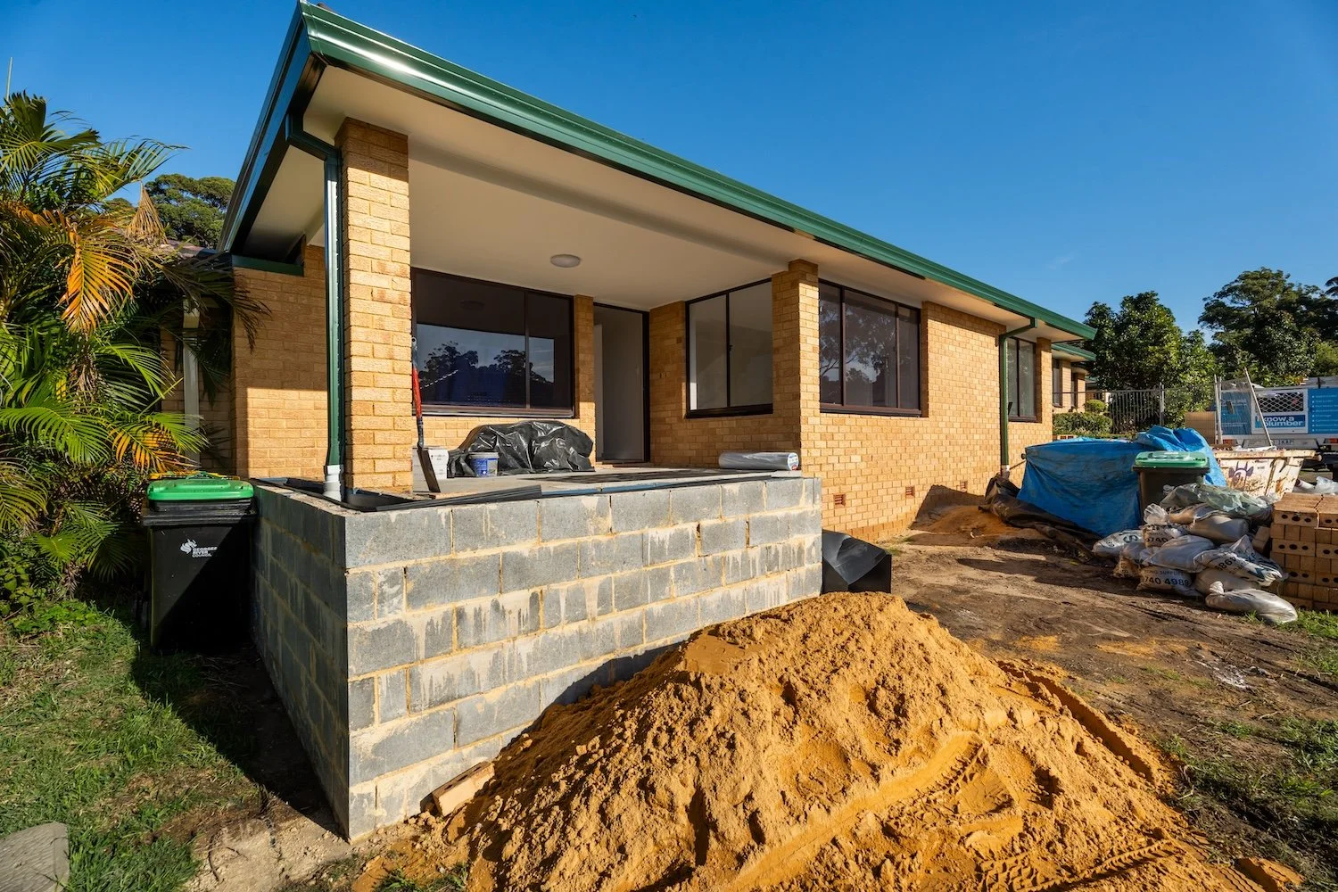 A house in South Australia with a brick exterior, large windows, and a green metal roof. There is a pile of dirt in the foreground and construction materials around the site. Insurance Builders