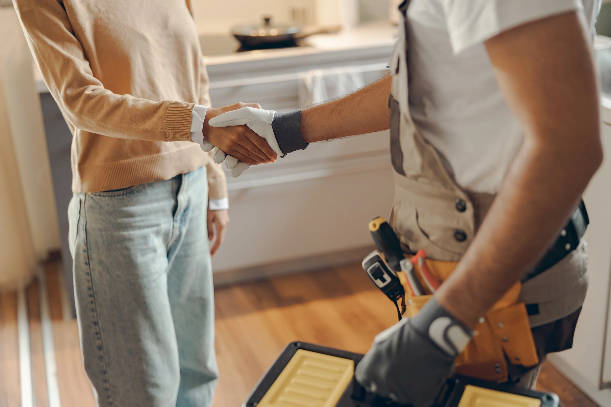A person wearing a beige sweater shaking hands with a person dressed as a construction worker with a tool belt, in a room with wooden floors and white cabinets.