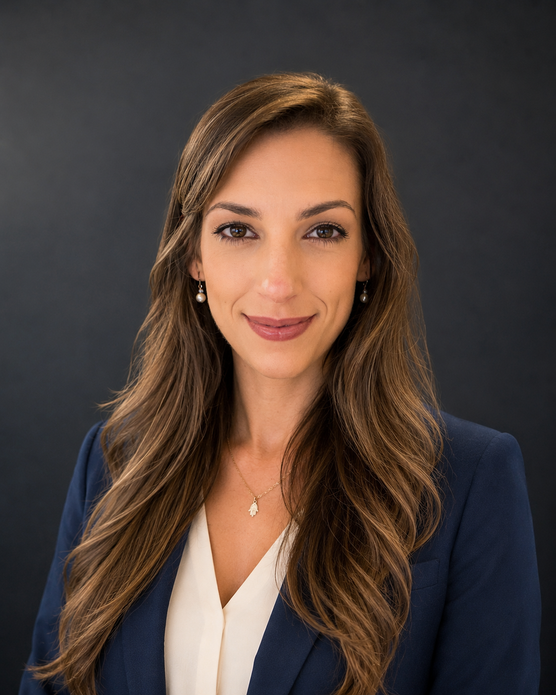 A professional woman with long brown hair wearing a navy blazer, cream blouse, pearl earrings, and a gold necklace with a hand pendant, smiling against a dark gray background.