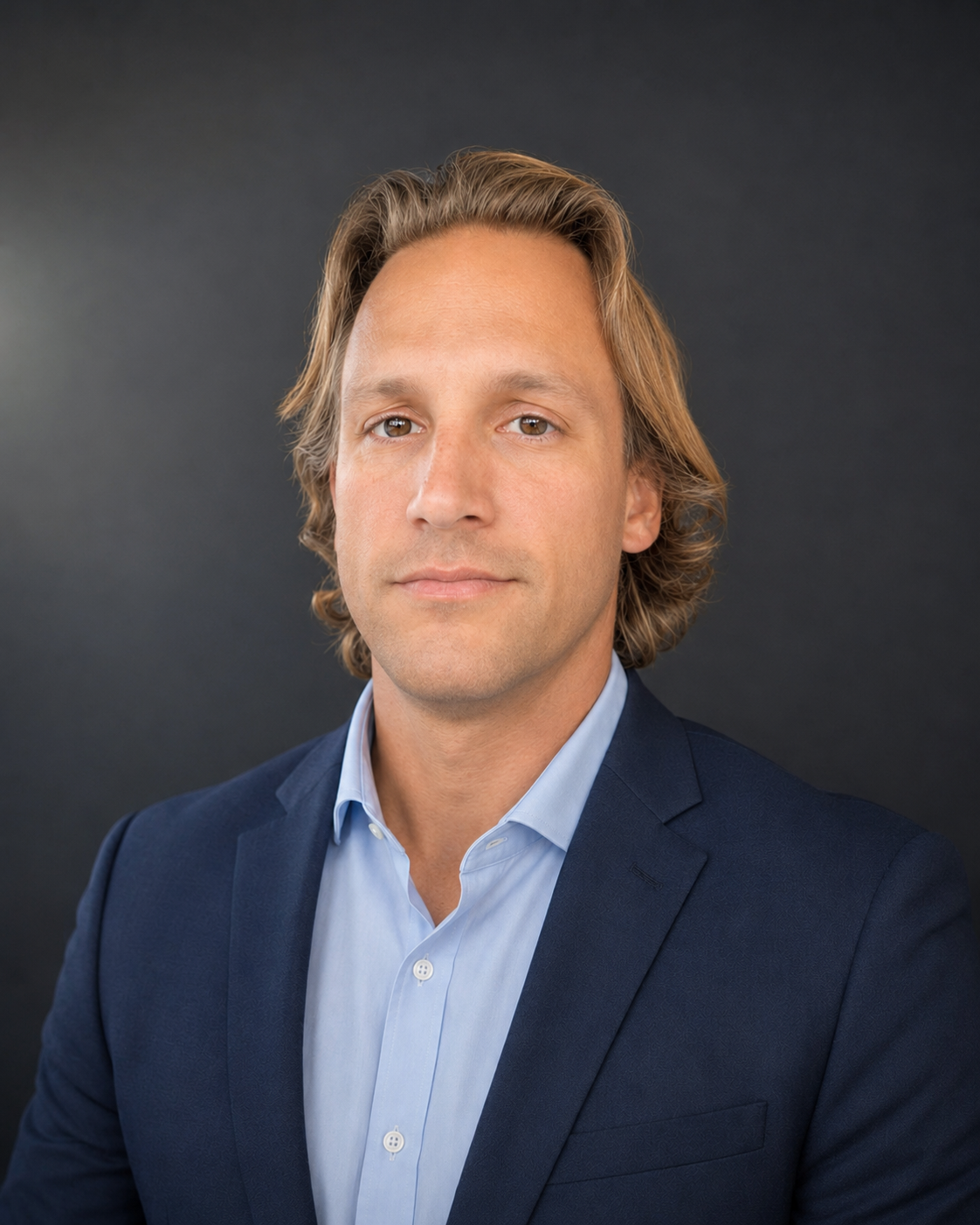 Headshot of a man with shoulder-length light brown hair, wearing a blue suit jacket and light blue dress shirt, against a dark gray background.