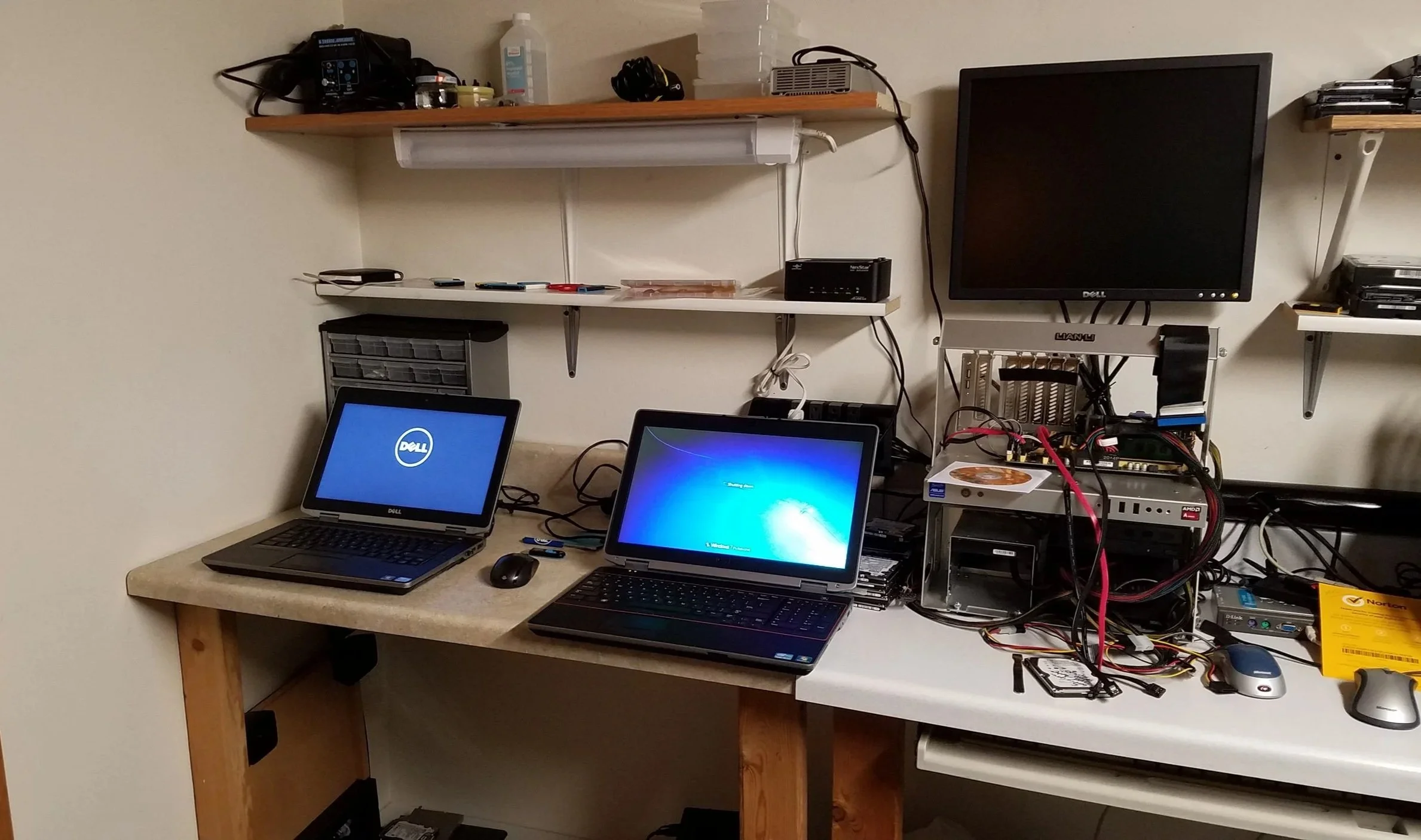 A cluttered workspace with two laptops on a wooden desk, one displaying a Dell logo and the other starting Windows, a monitor off, and a variety of electronic components, wires, and tools on shelves and the right side of the desk.